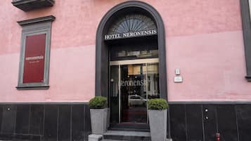 Ein Hotel Eingang mit einem Schild, auf dem 'HOTEL NERONENSIS' steht, einer Glastür und zwei Blumentöpfen auf den Stufen.
