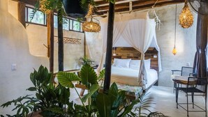 Ein gemütliches Zimmer mit Bett, Sessel und Tisch. Pflanzen und Pendelleuchten sind vorhanden.