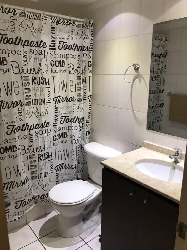 A bathroom with a white toilet, a sink, and a shower curtain with words like 'toothpaste', 'shampoo', and 'razor'.