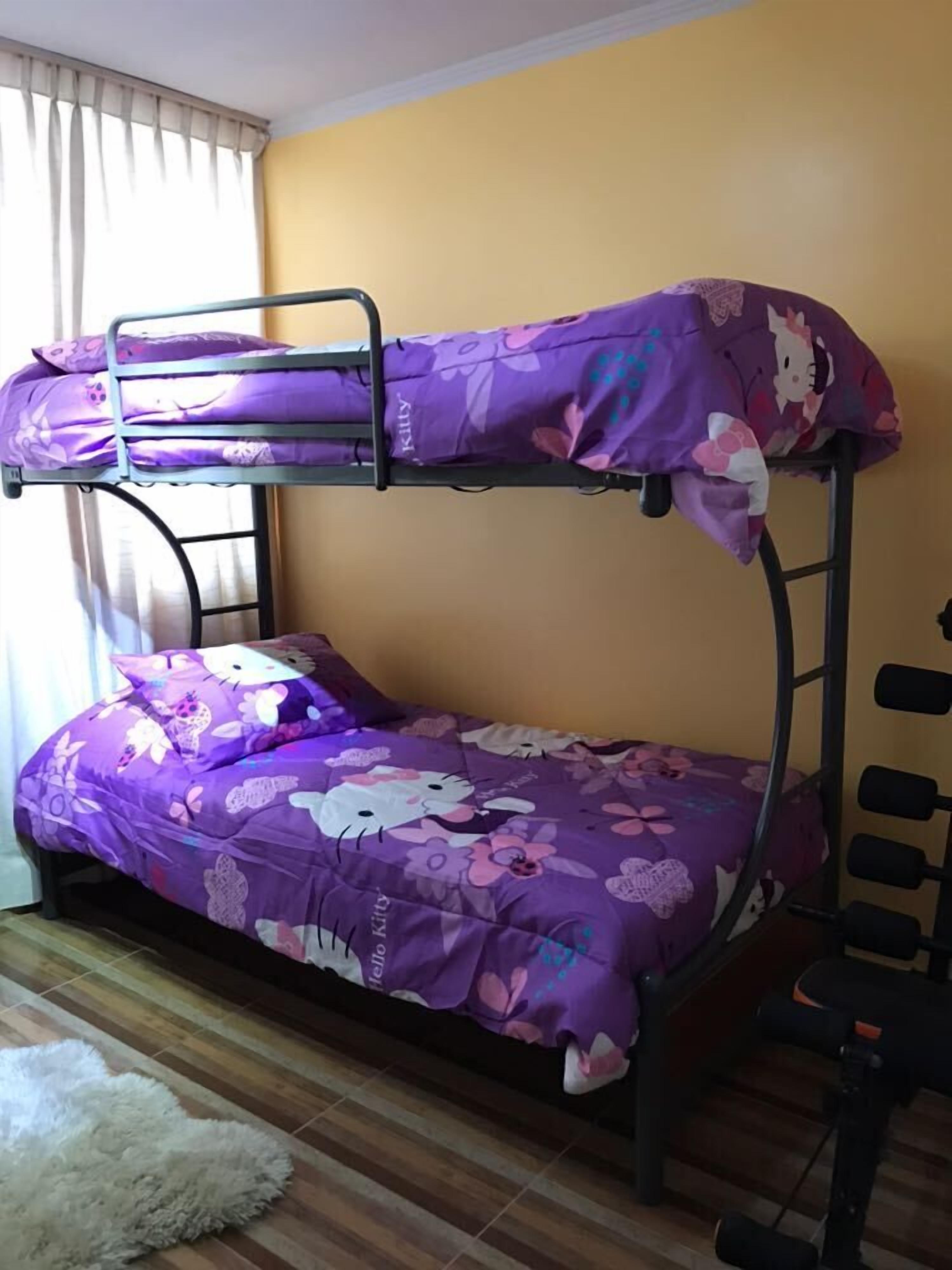 Un lit superposé avec une literie violette ornée de motifs Hello Kitty, un tapis blanc et moelleux et une fenêtre avec des rideaux.