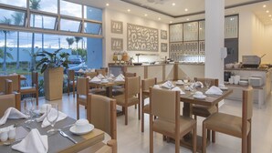 A modern dining area with tables set for service, a kitchen in the background, and large windows offering a view of palm trees and a cityscape. - whala!urban punta cana (Punta Cana)