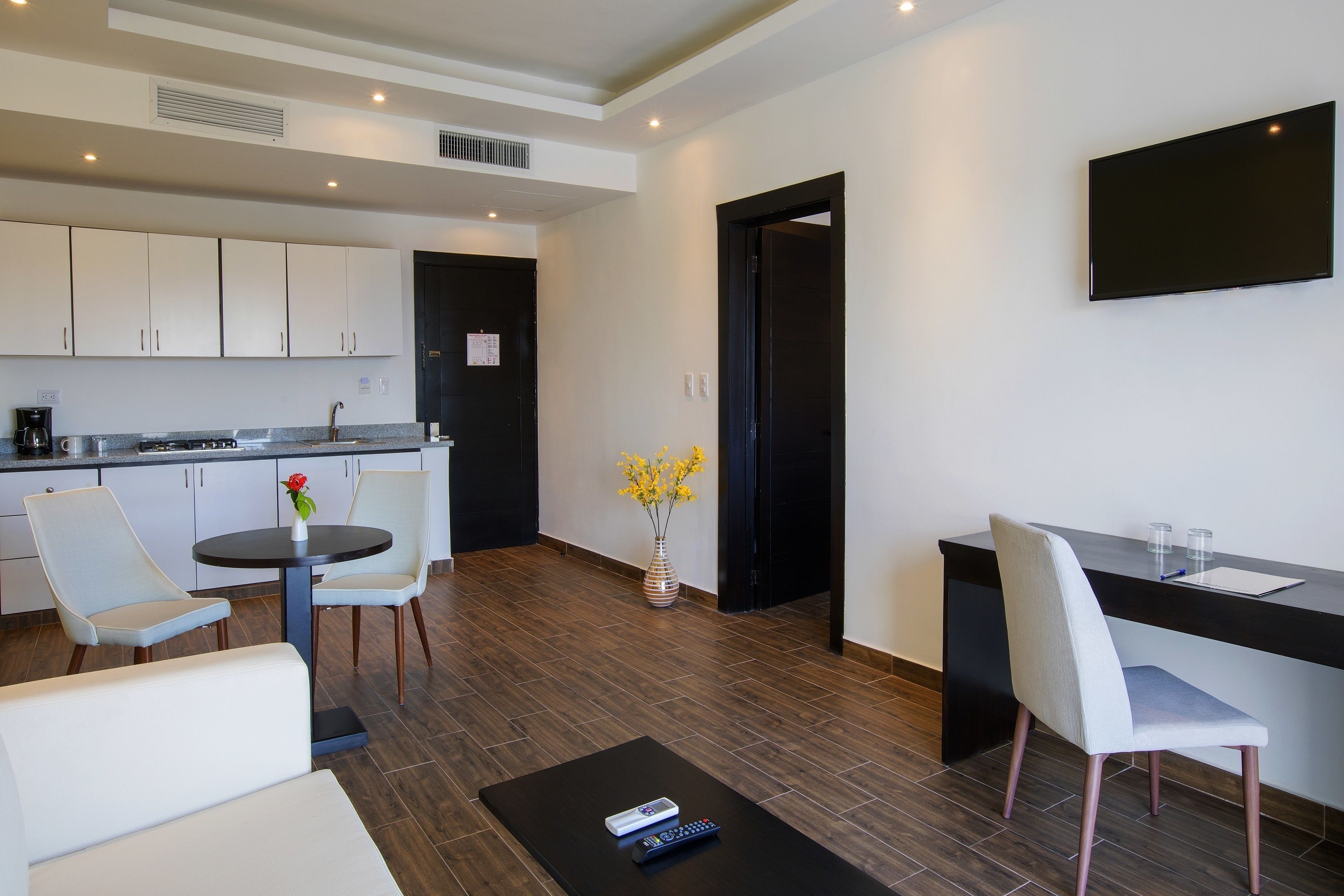 A modern kitchen with white cabinets, a dining area with a round table and chairs, a small coffee table with remote controls, and a vase with yellow flowers.
