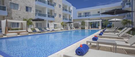 Una piscina con sillas de descanso y sombrillas, rodeada de edificios de varios pisos con balcones.