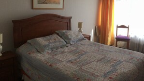 Une chambre à coucher avec un lit en bois, une literie à motifs floraux, une table de chevet, une lampe, une chaise et un tableau accroché au mur.