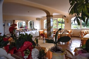 Un comedor con mesas, sillas, plantas en macetas y ventanas grandes.