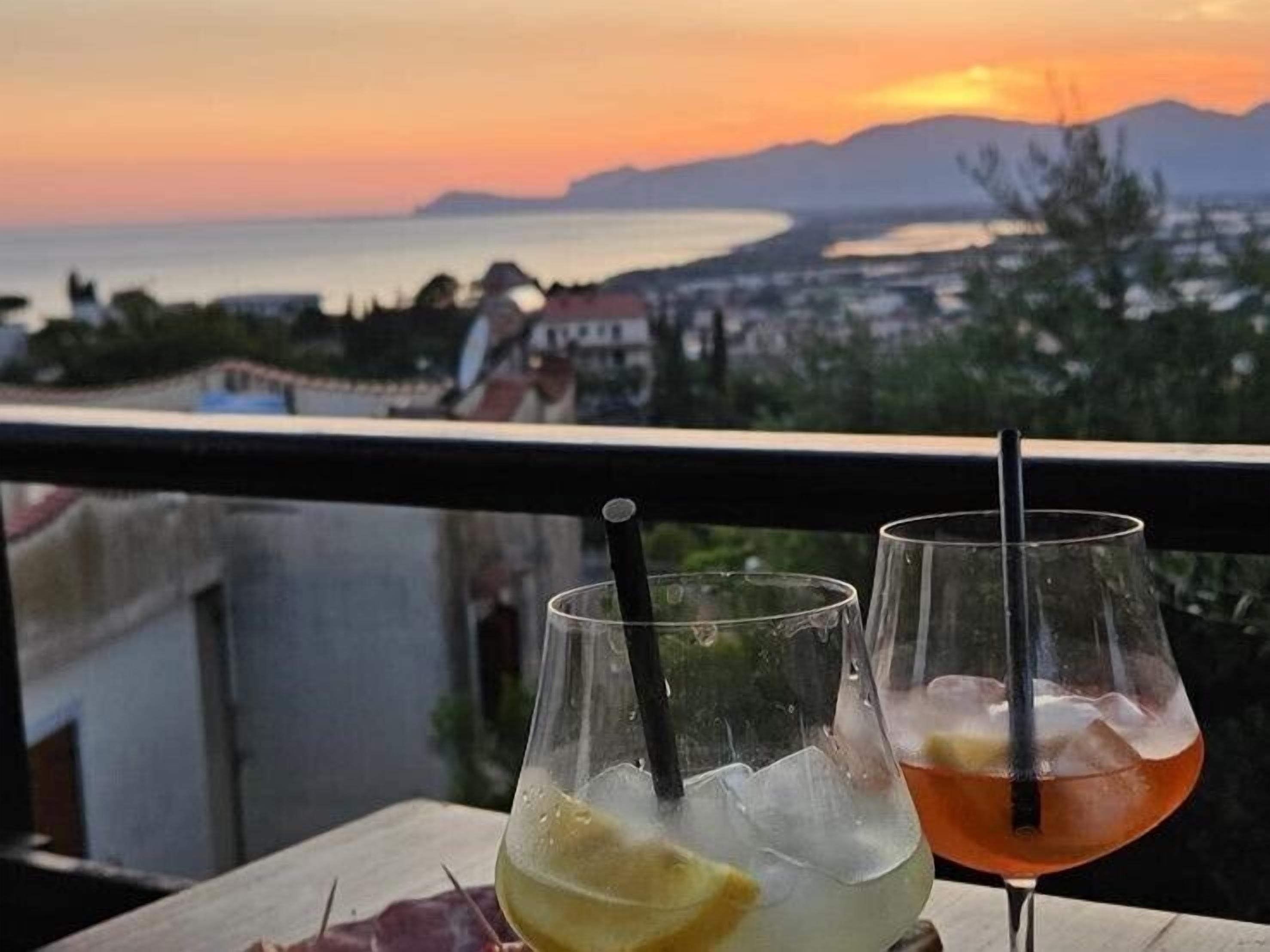 Two glasses of beverages on a balcony with a view of the sea and mountains.