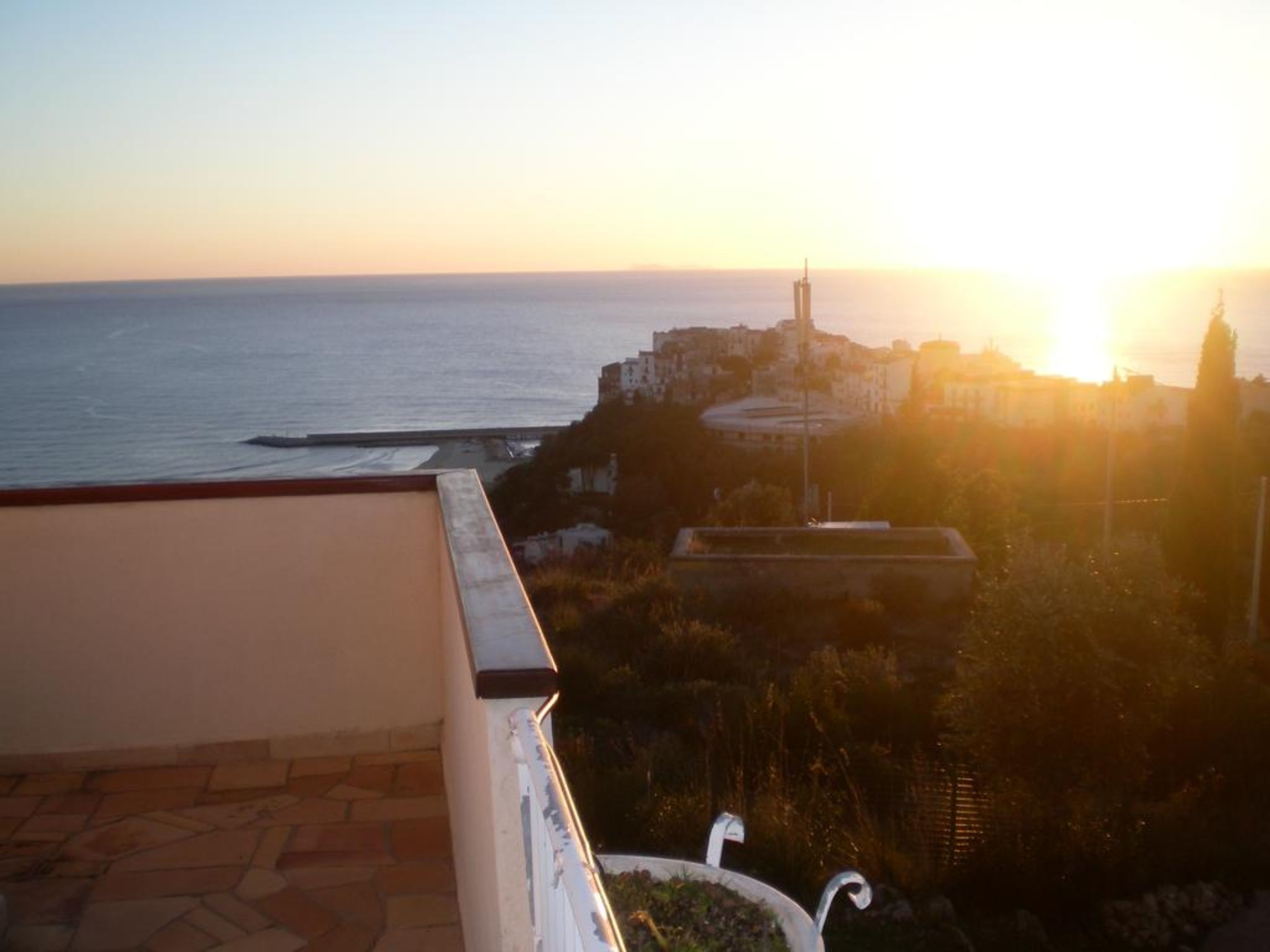 Vista costera con atardecer, un muelle y edificios al fondo.