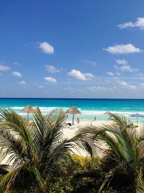 A beach with clear turquoise water, palm trees, and beach umbrellas. - Apartamento ZH 607 by Sinbad (Cancun)