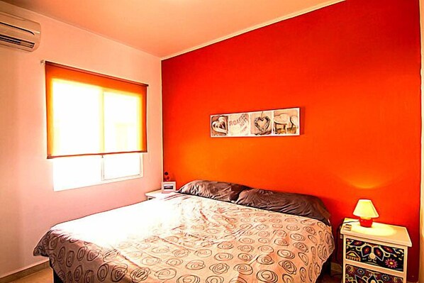A bedroom with a bed, a nightstand with a lamp, a window with blinds, and a wall with a framed picture.