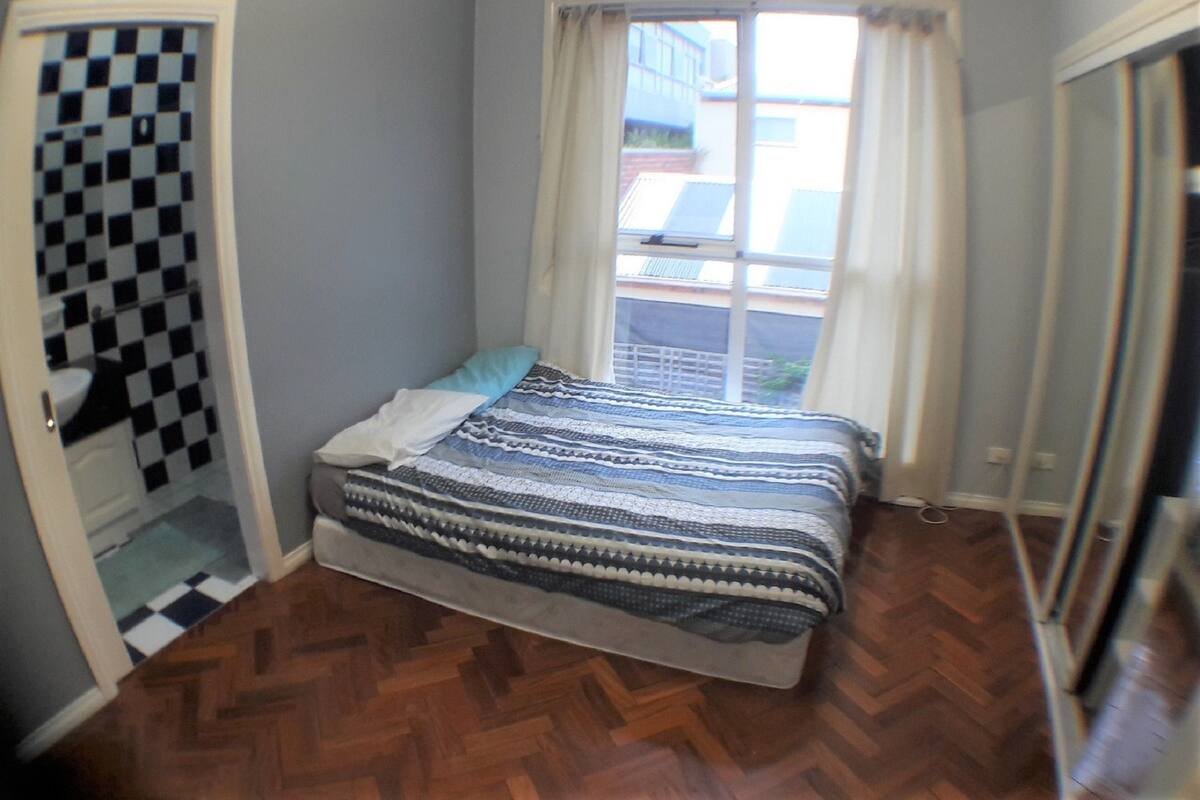 Een slaapkamer met een zwart-wit geruit vloer, een bed met een geborduurde deken, een raam met gordijnen en een deur die leidt naar een badkamer met een geruit vloer.