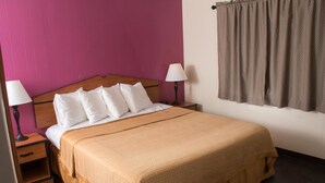 A neatly made bed with white pillows and a beige bedspread, flanked by two bedside lamps and a nightstand with a drawer. - Budget Inn (Caldwell)