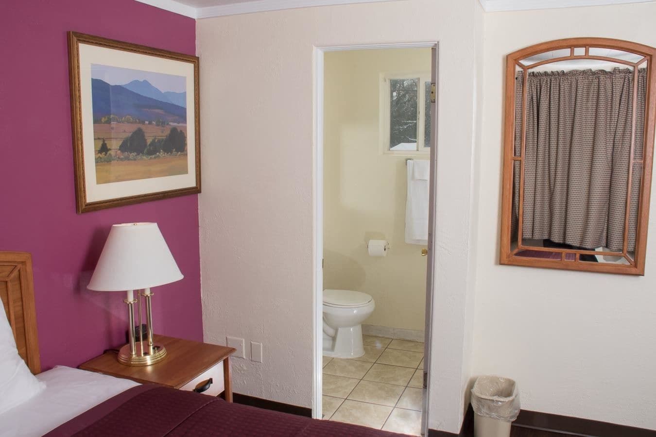 Une chambre à coucher avec un lit, une table de chevet avec une lampe, un tableau encadré au mur et une vue sur la salle de bain par une porte ouverte.