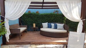 A covered outdoor seating area with wicker furniture and a cushioned bench, surrounded by a blue wall and greenery.