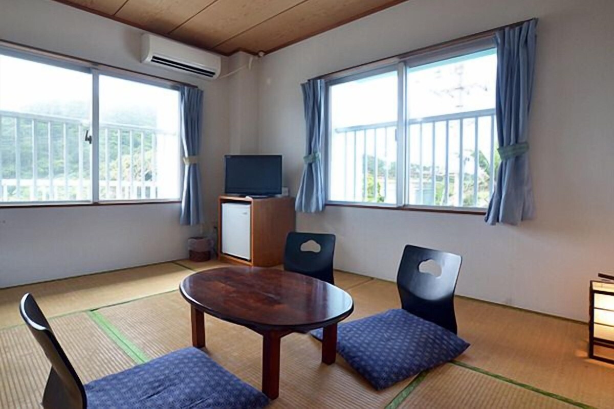 A room with tatami flooring, a wooden table, two chairs, a television on a cabinet, and large windows with curtains.