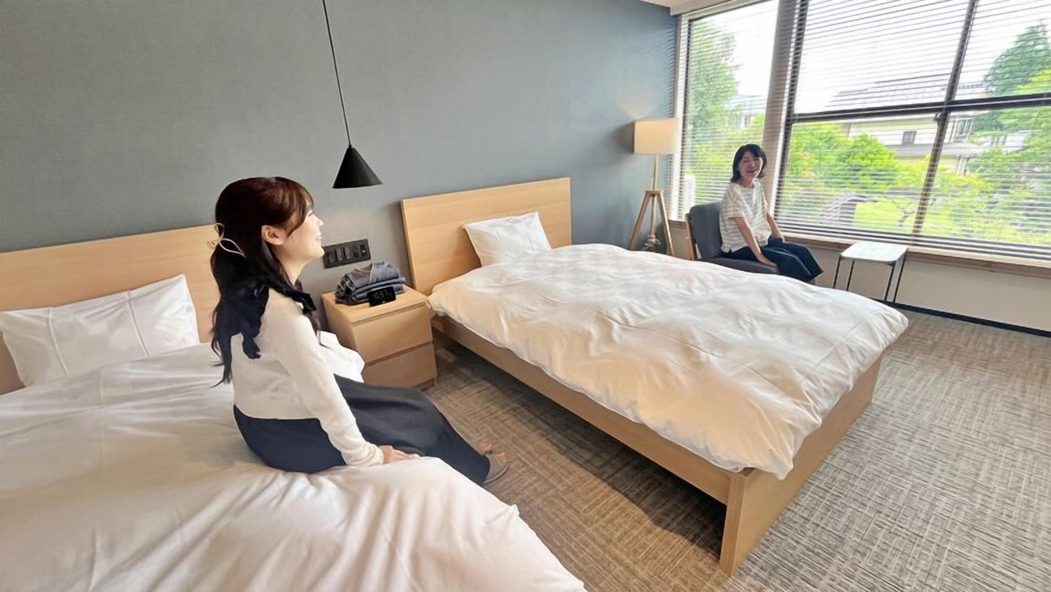 A hotel room with two beds, a nightstand, a lamp, and a window with a view of greenery.
