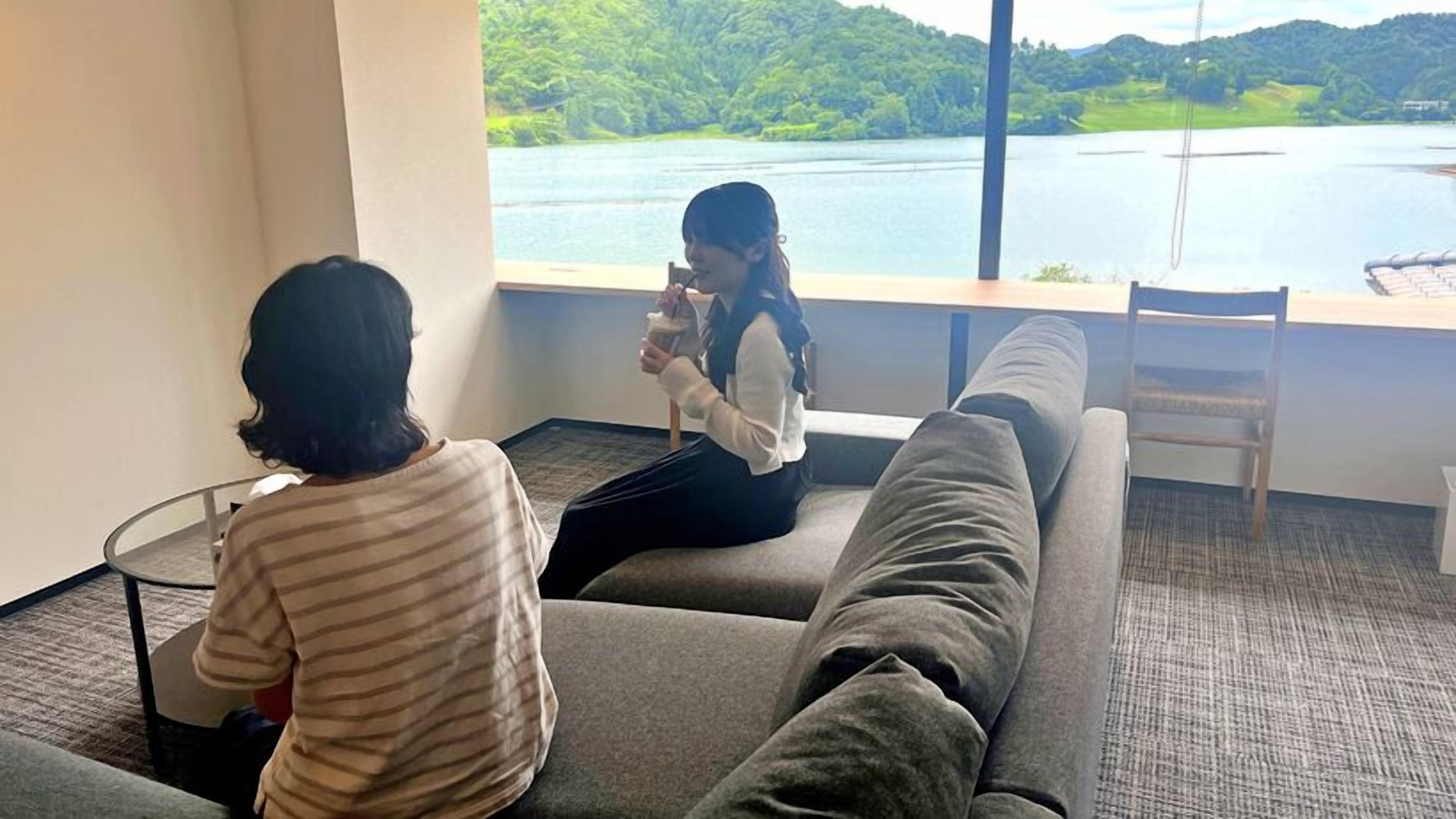 Two people sitting on a couch facing a window with a view of a lake and hills.
