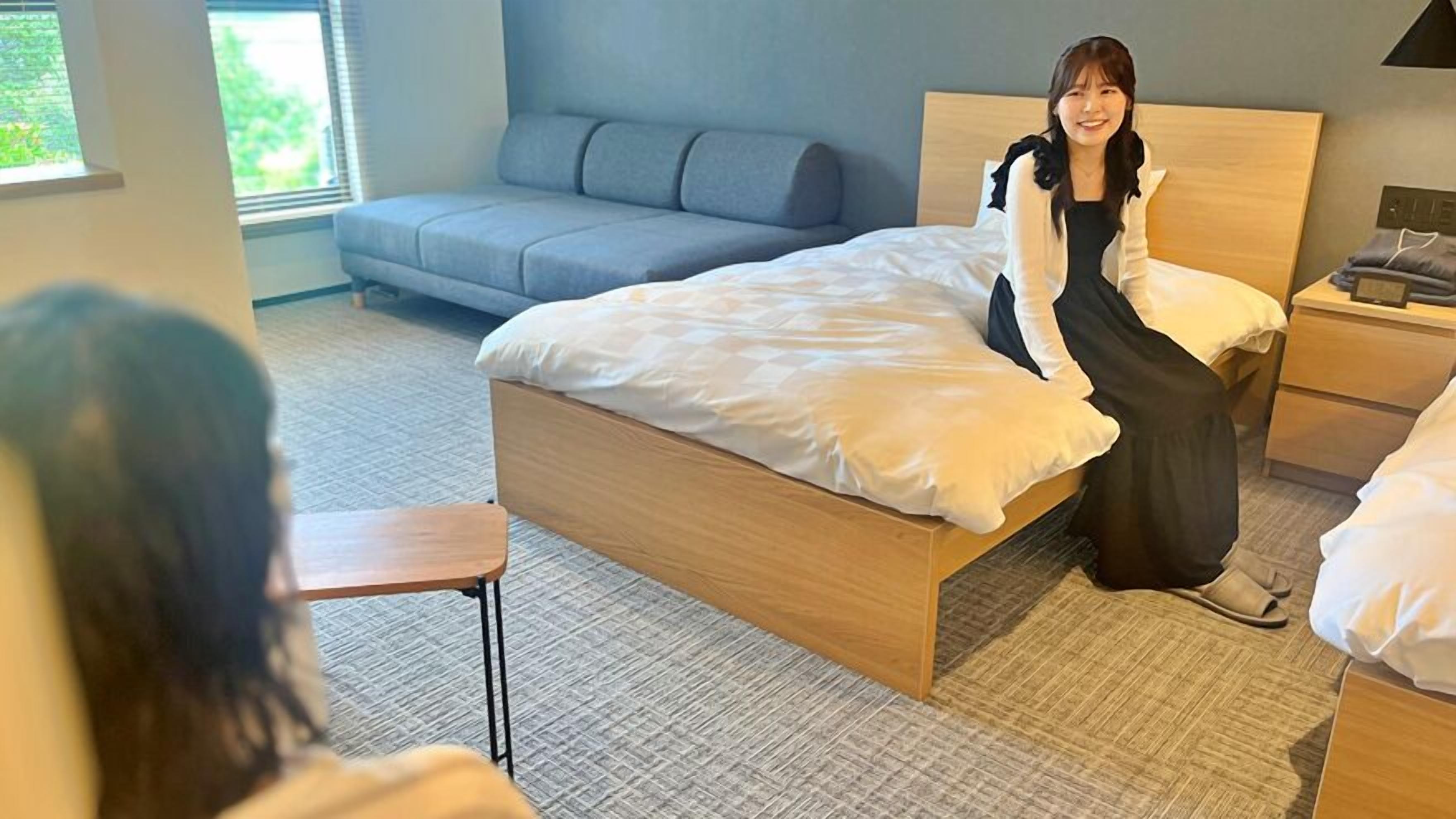 A hotel room with a bed, a bedside table, a nightstand, a sofa, and a window.