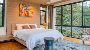 A modern bedroom with a large bed, a painting on the wall, and a view of greenery through large windows. - Moin Lakeside Pool Villa ChiangMai (Hang Dong)