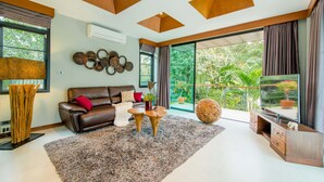 A modern living room with a brown leather sofa, a large flat-screen TV, and a wooden coffee table. There is a large window with a view of greenery outside. - Moin Lakeside Pool Villa ChiangMai (Hang Dong)