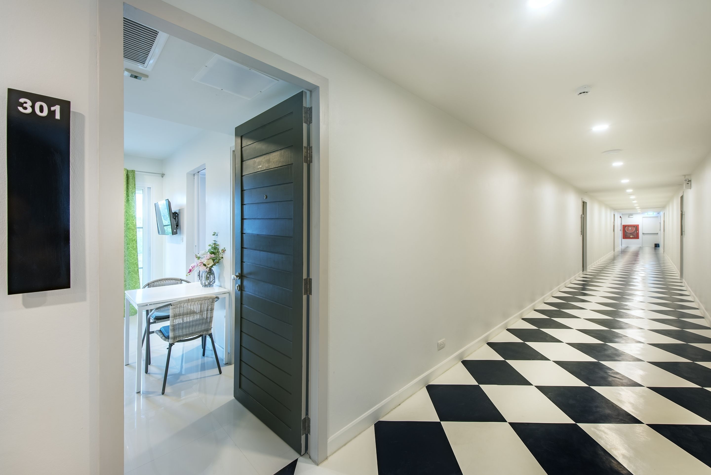 Un pasillo de hotel con suelo a cuadros blanco y negro, una puerta con la inscripción '301', y un área de comedor con una mesa y sillas.
