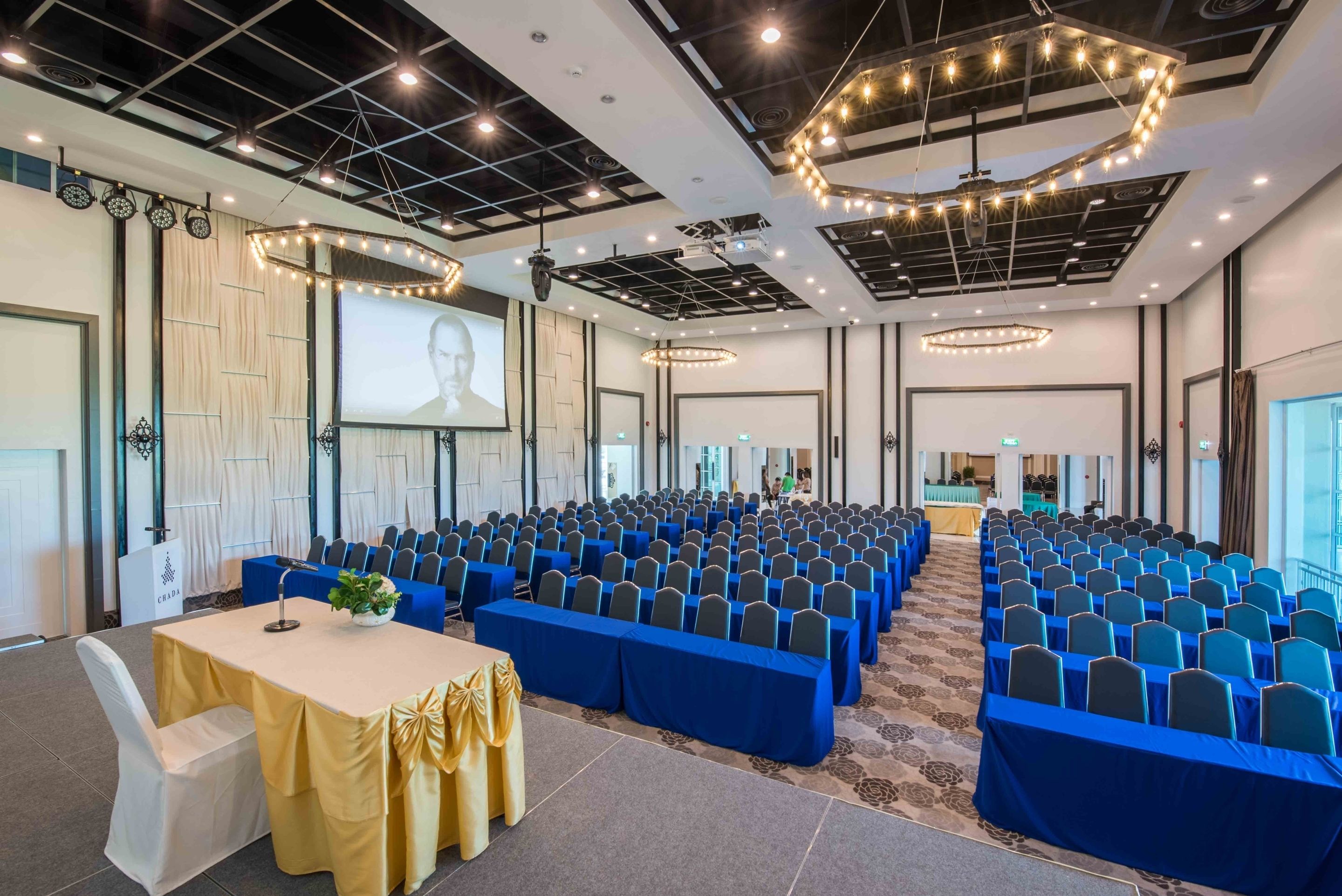 Uma sala de conferências com fileiras de cadeiras azuis, um palco com uma tela e um teto com luzes.