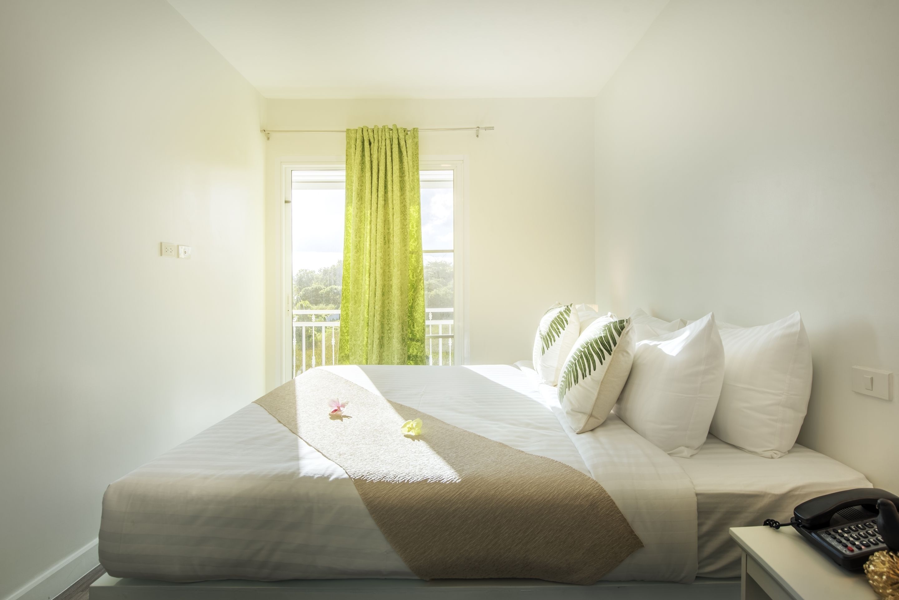 A neatly made bed with a white and beige bedspread, a green curtain, and a telephone on a bedside table.