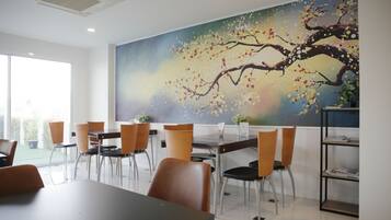 A modern dining area with a large mural of a tree on the wall, a long table with chairs, and a bookshelf.