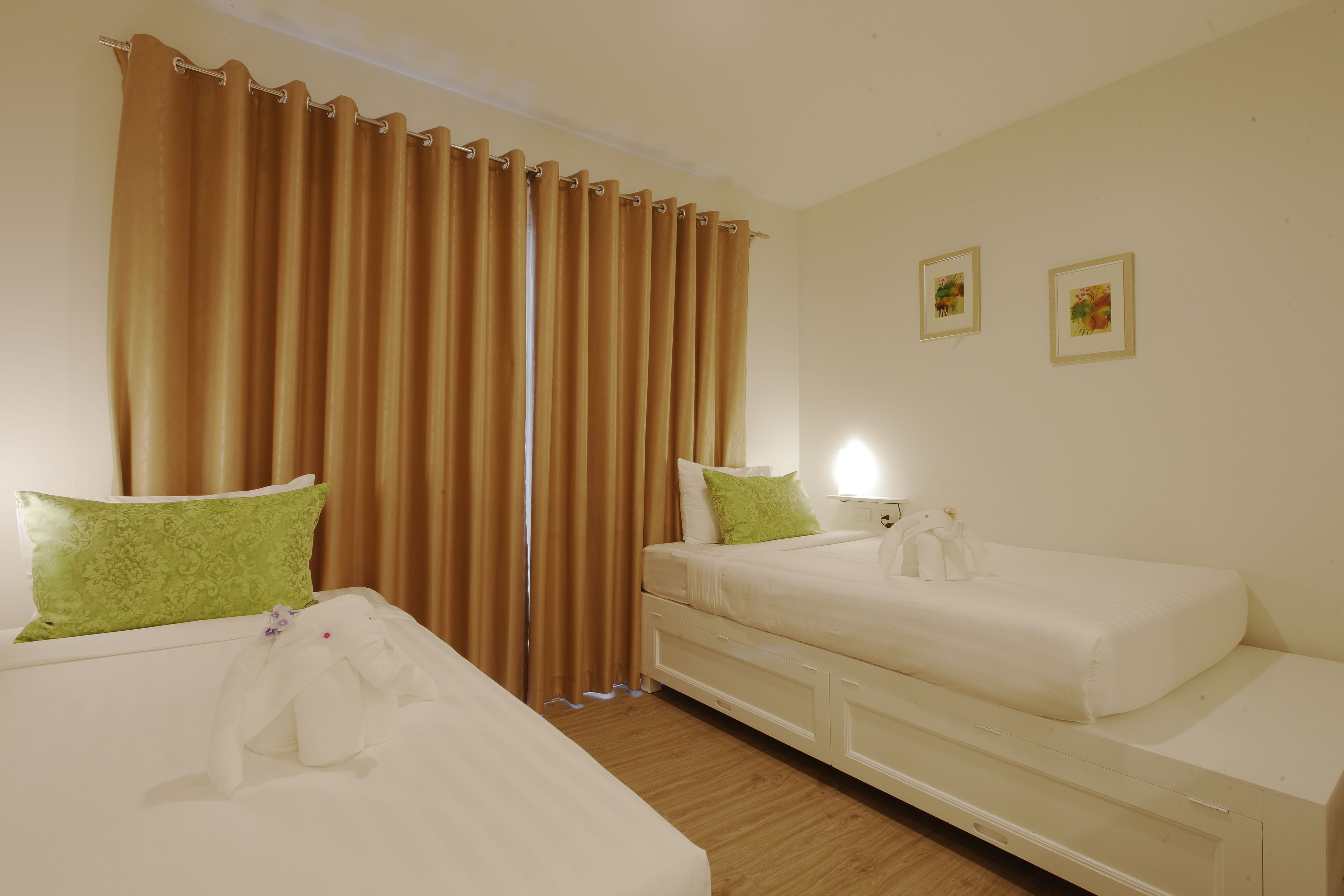 A hotel room with two beds, a large window with curtains, a green pillow, and a small table with a lamp.