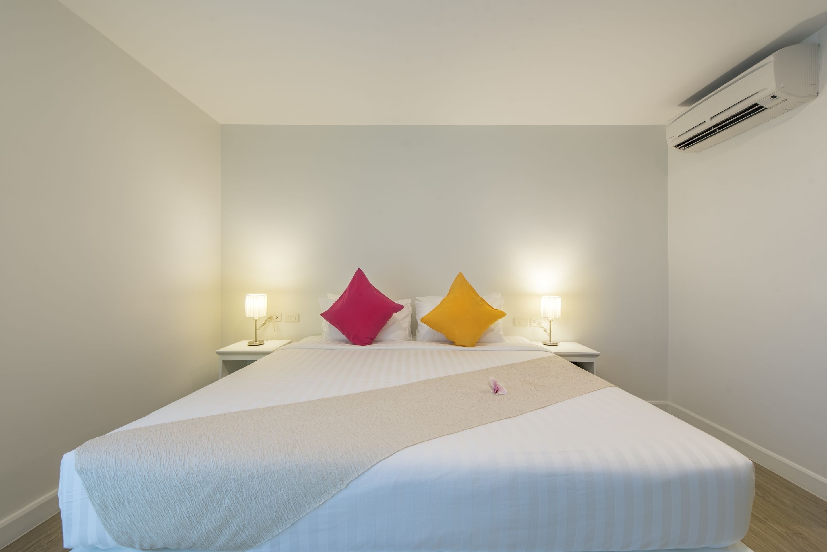 A neatly made bed with white bedding and two pillows, one pink and one yellow, in a simple room with a white wall and an air conditioning unit.