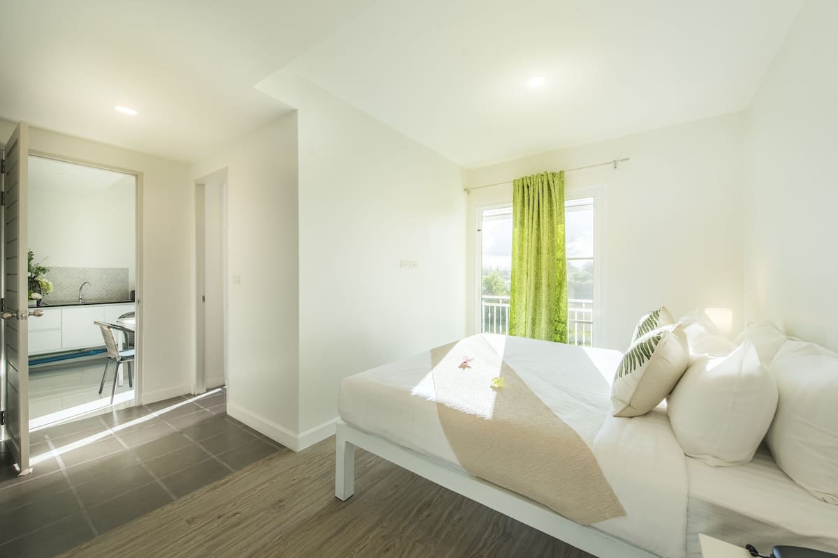 Quarto de hotel moderno com uma cama, uma janela com cortinas verdes, vista para o exterior e uma cozinha compacta no espaço adjacente.