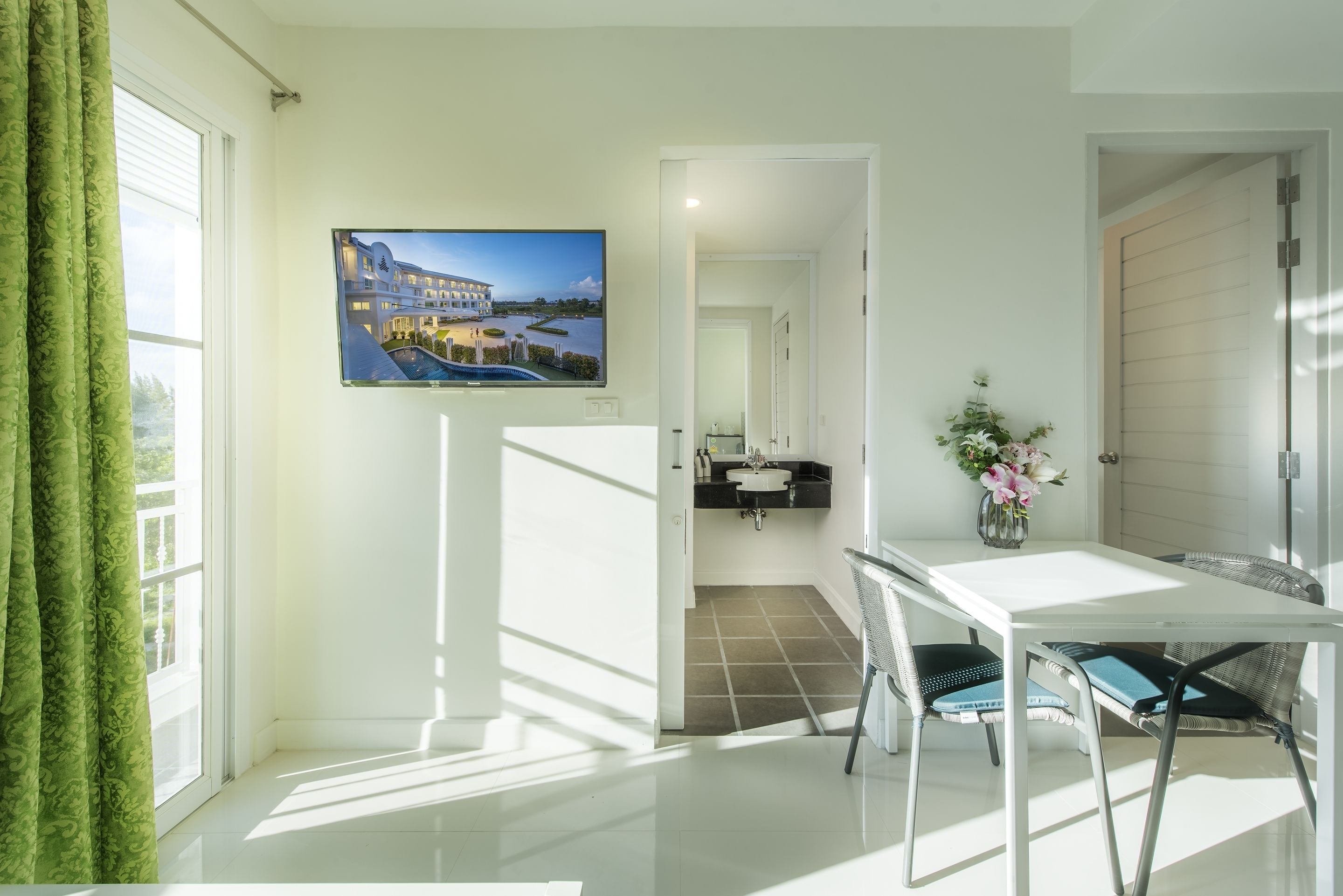 A bright room with a TV displaying a resort, a white table with chairs, and a view of a pool through the window.