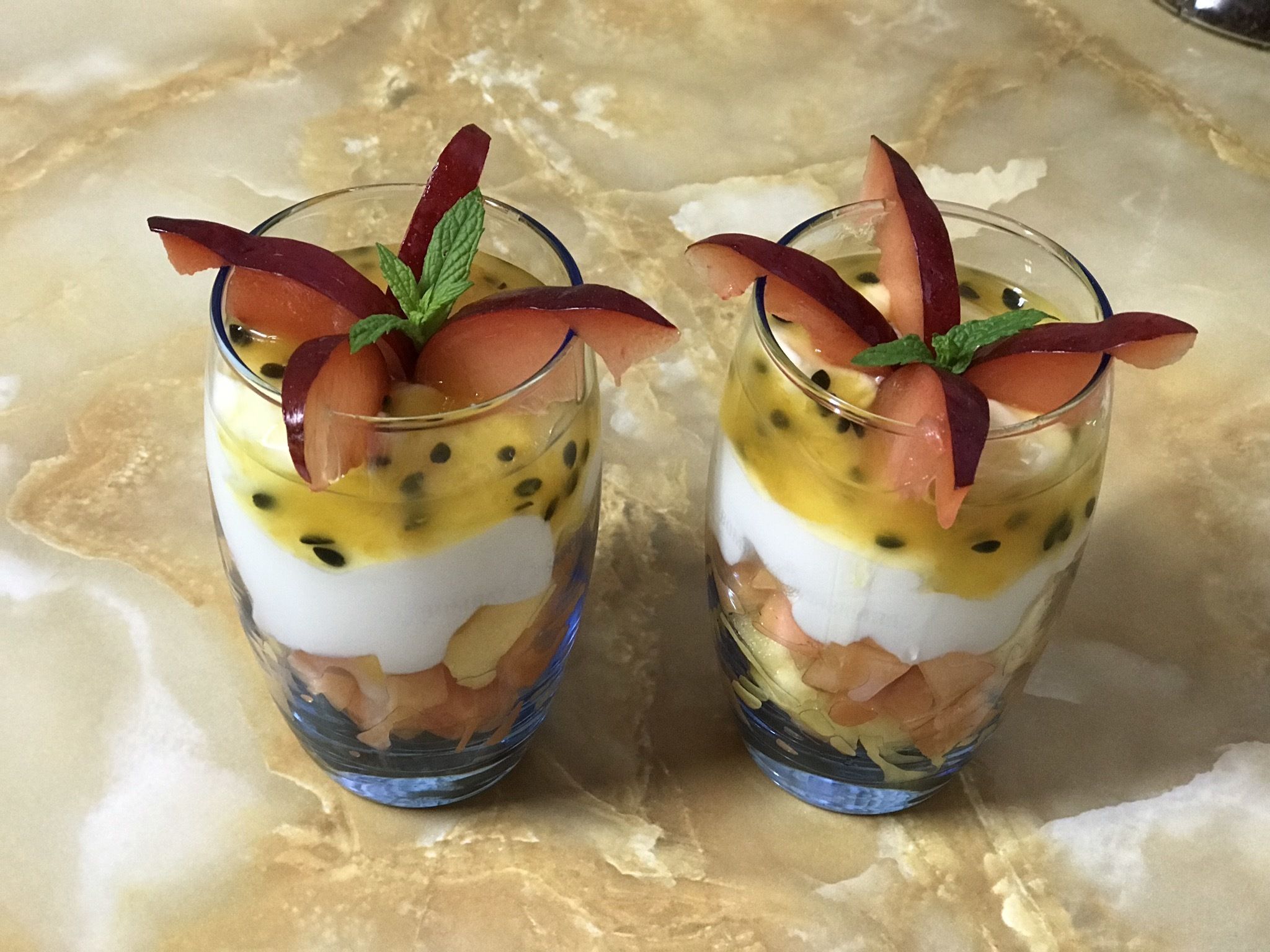 Deux coupes de dessert aux fruits en couches, garnies de feuilles de menthe et de tranches de fruits rouges, posées sur une surface en marbre.
