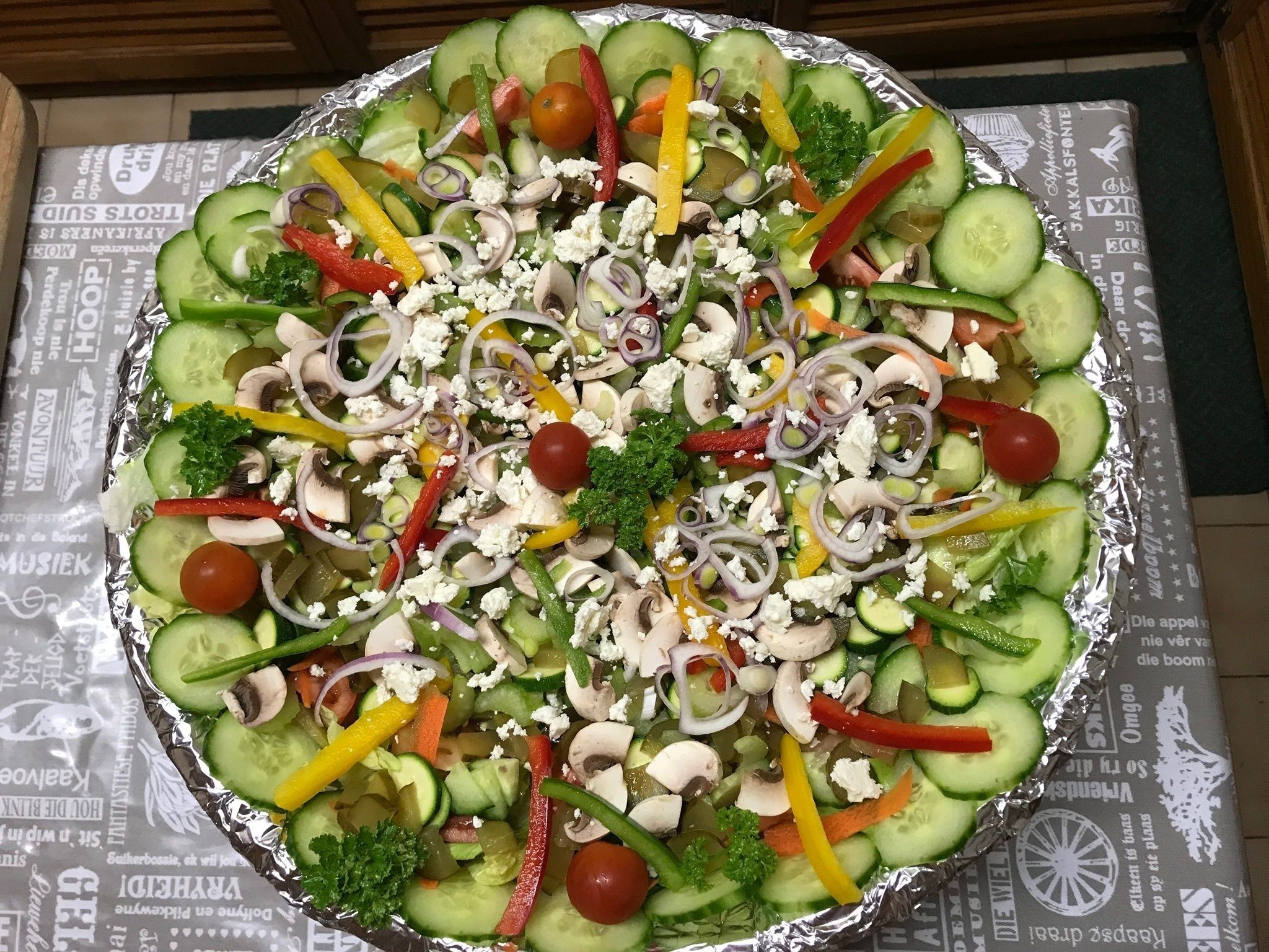 A large, round, foil-covered salad with various vegetables and feta cheese.