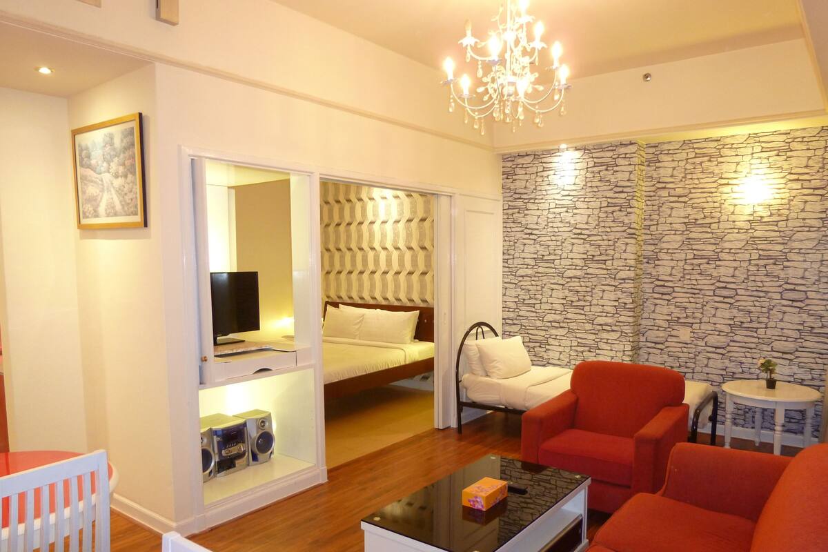 A compact living space with a bed, a television, a red armchair, a small table, and a chandelier.