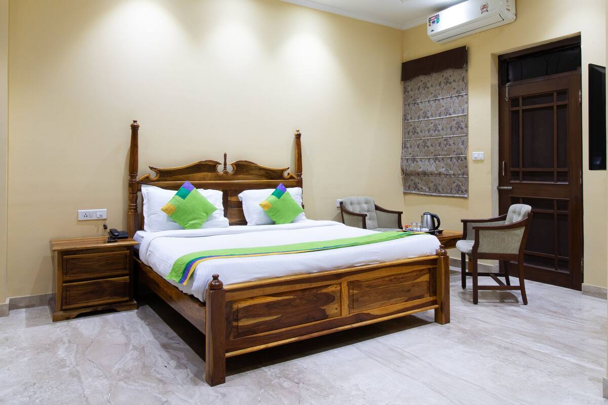A wooden four-poster bed with white bedding and colorful pillows, a bedside table with a phone, a chair, and a window with a floral blind.