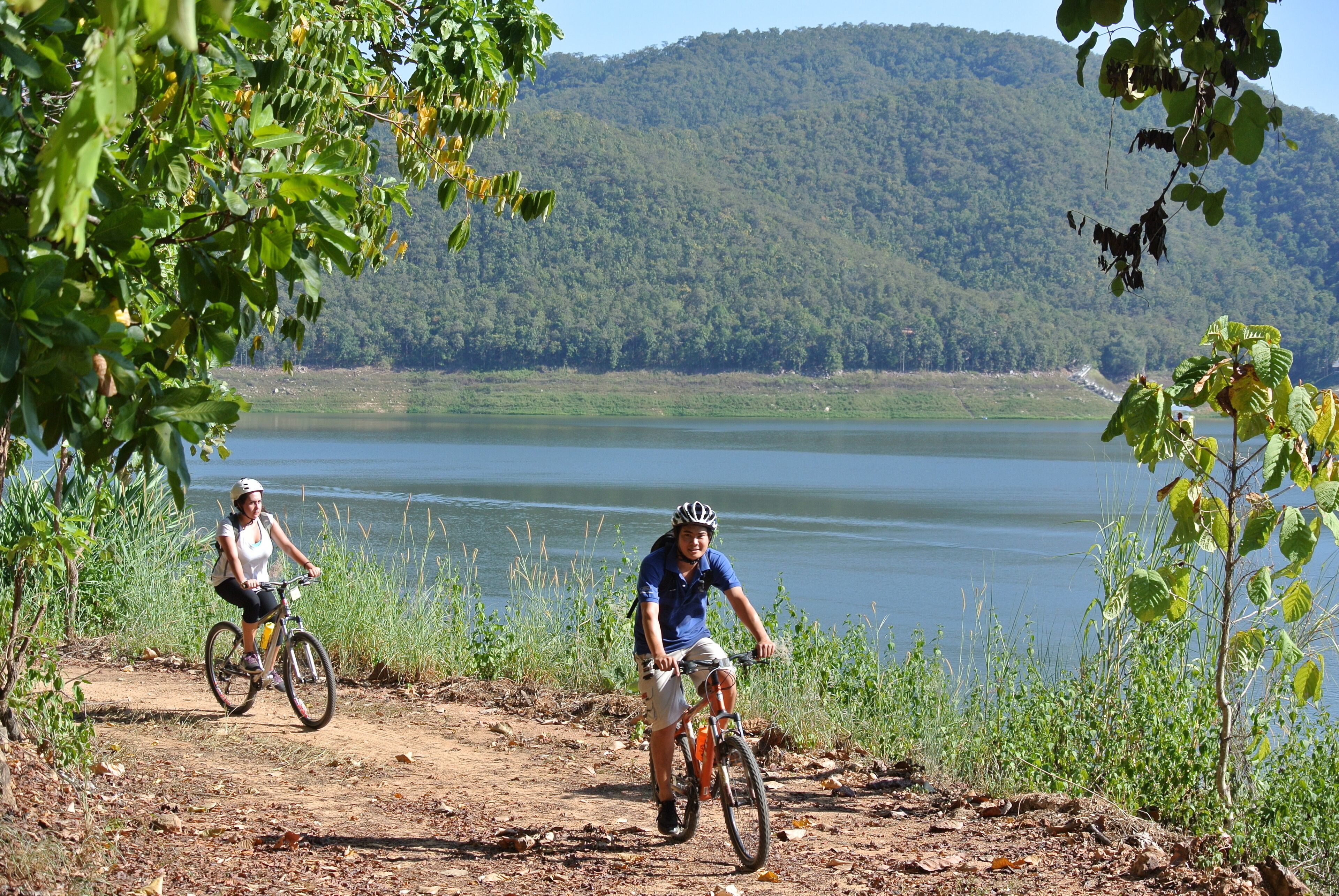 Dois ciclistas numa trilha de terra com um lago e colinas arborizadas ao fundo.