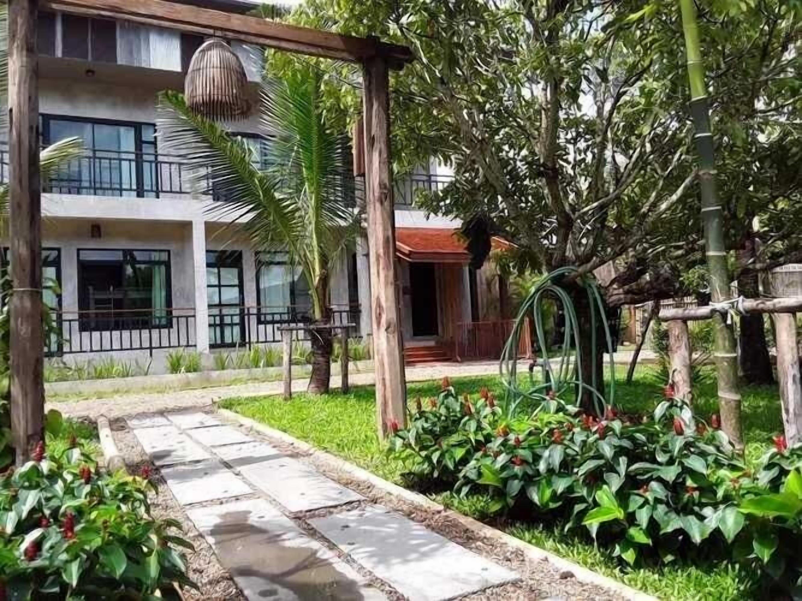 A pathway leading to a building with a balcony, surrounded by lush greenery and a garden area with plants and a hanging light fixture.
