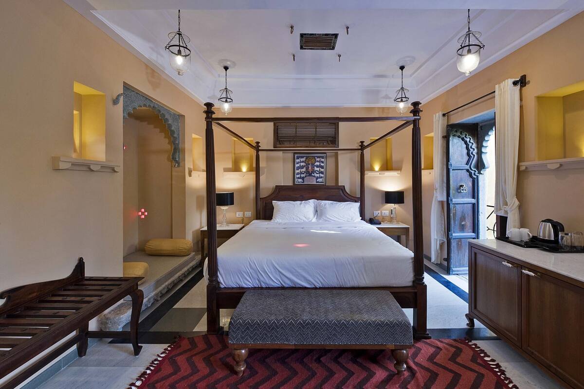 A four-poster bed with a wooden frame, a bench at the foot of the bed, and a red and black patterned rug on the floor.