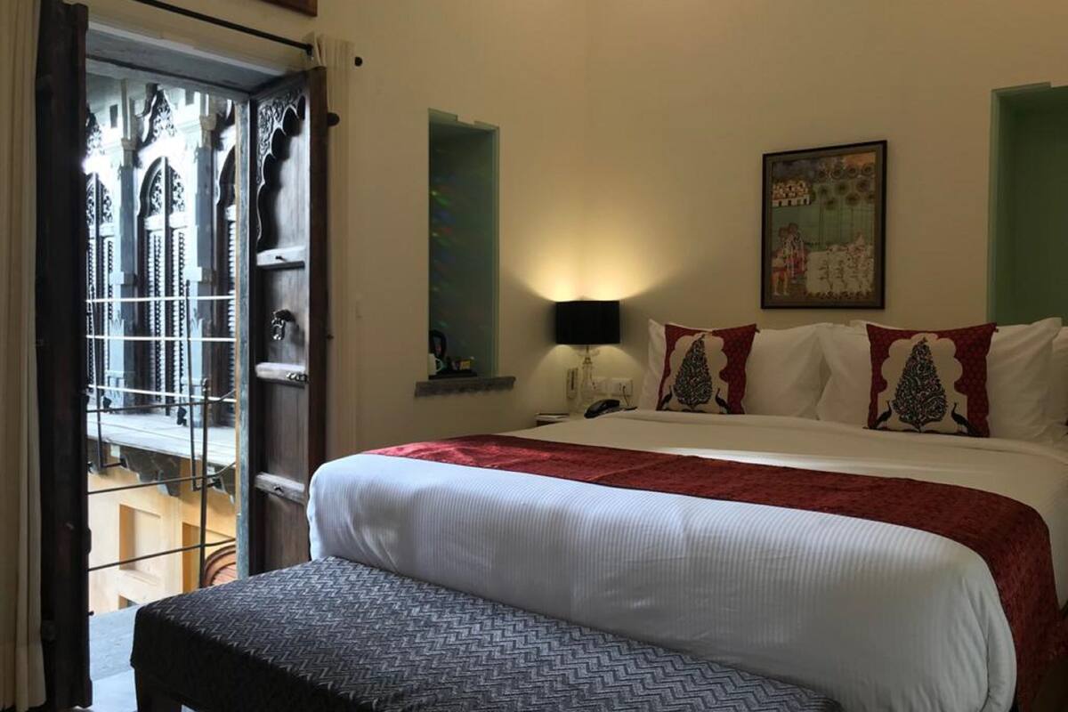 A hotel room with a large bed, a bedside table with a lamp, a painting on the wall, and a view through a window with curtains.
