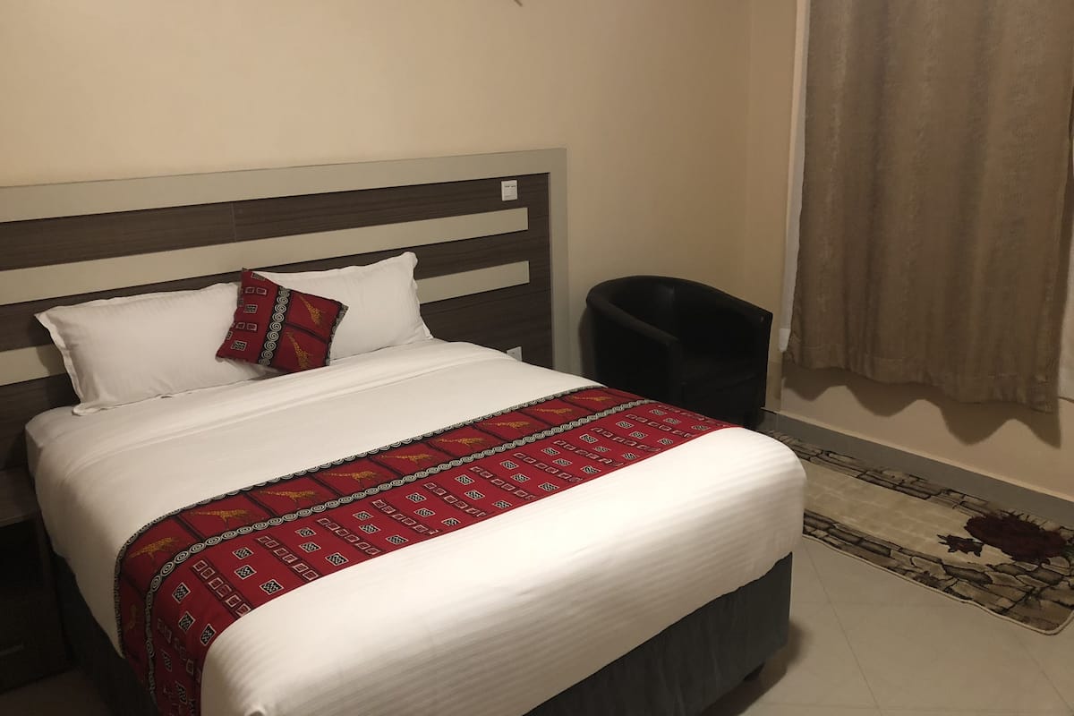 Un lit avec une couvre-lit à motifs, un oreiller, une lampe fixée au mur, une poubelle noire et un tapis à motifs.