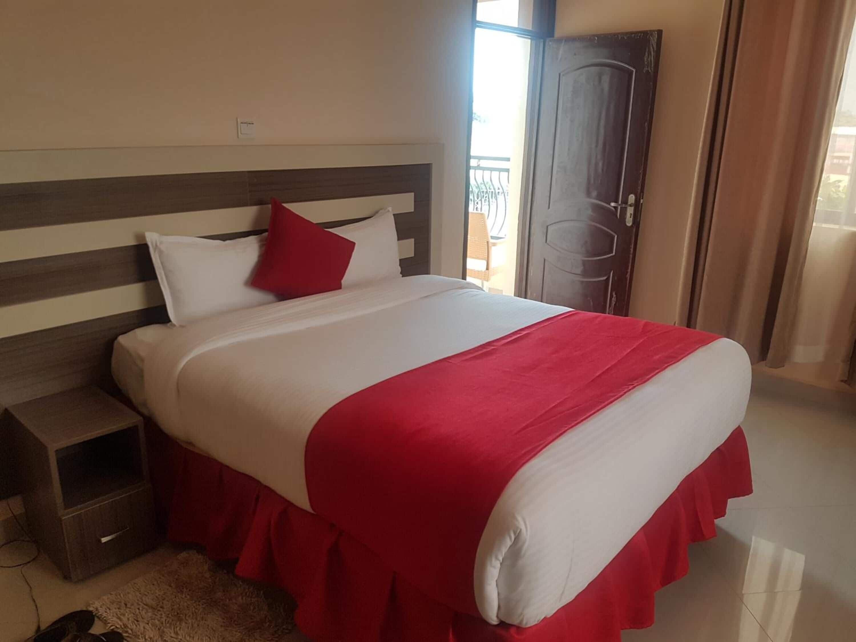 A hotel room with a bed, a nightstand, a door, and a view of the outside.