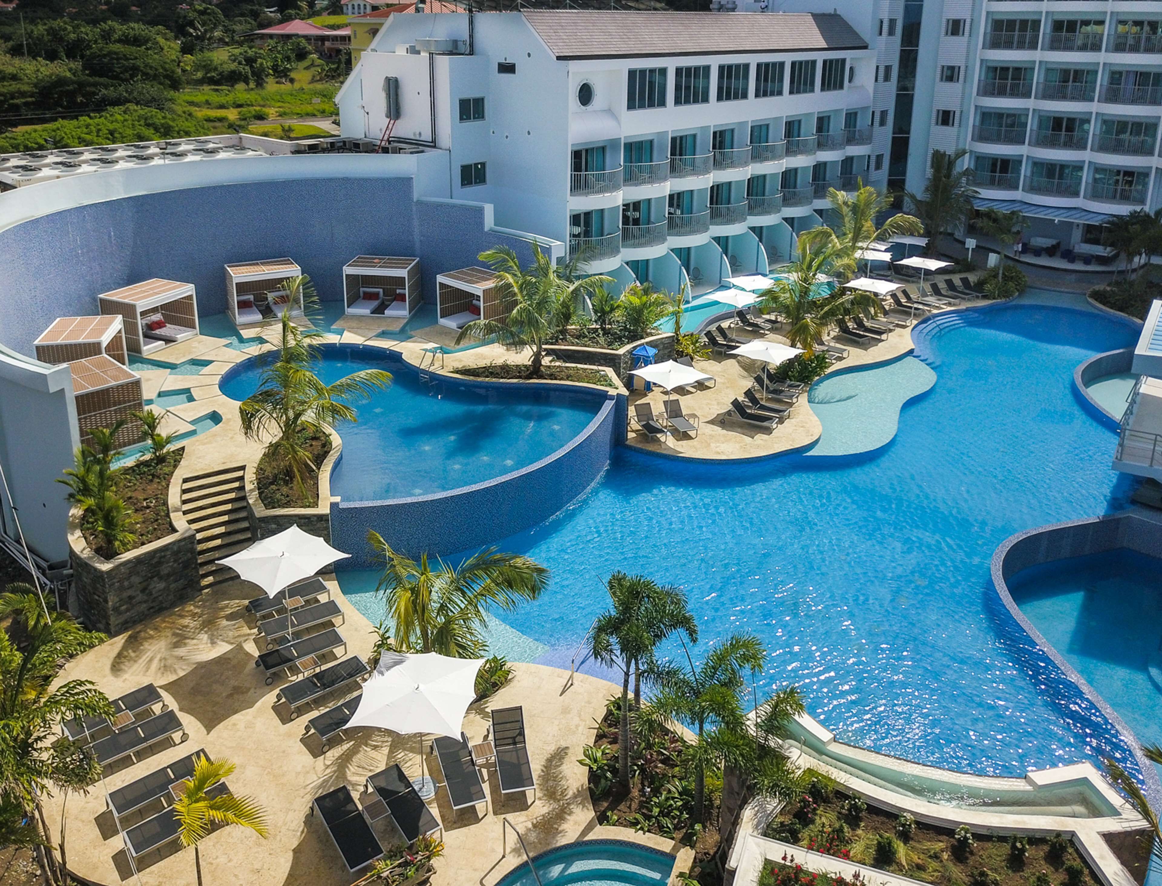 A resort with a large, curved swimming pool surrounded by lounge chairs and umbrellas, with a building in the background.