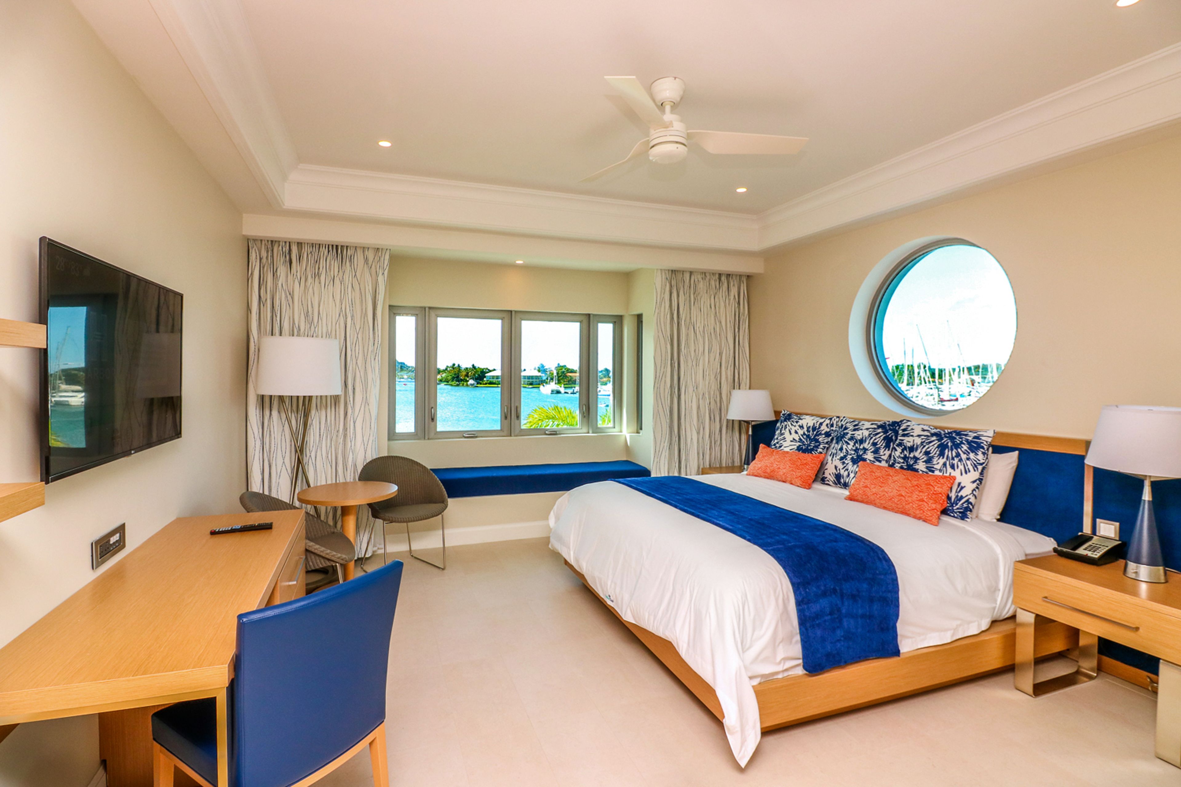 A hotel room with a bed, a desk, a chair, a TV, a window with a view, and a ceiling fan.