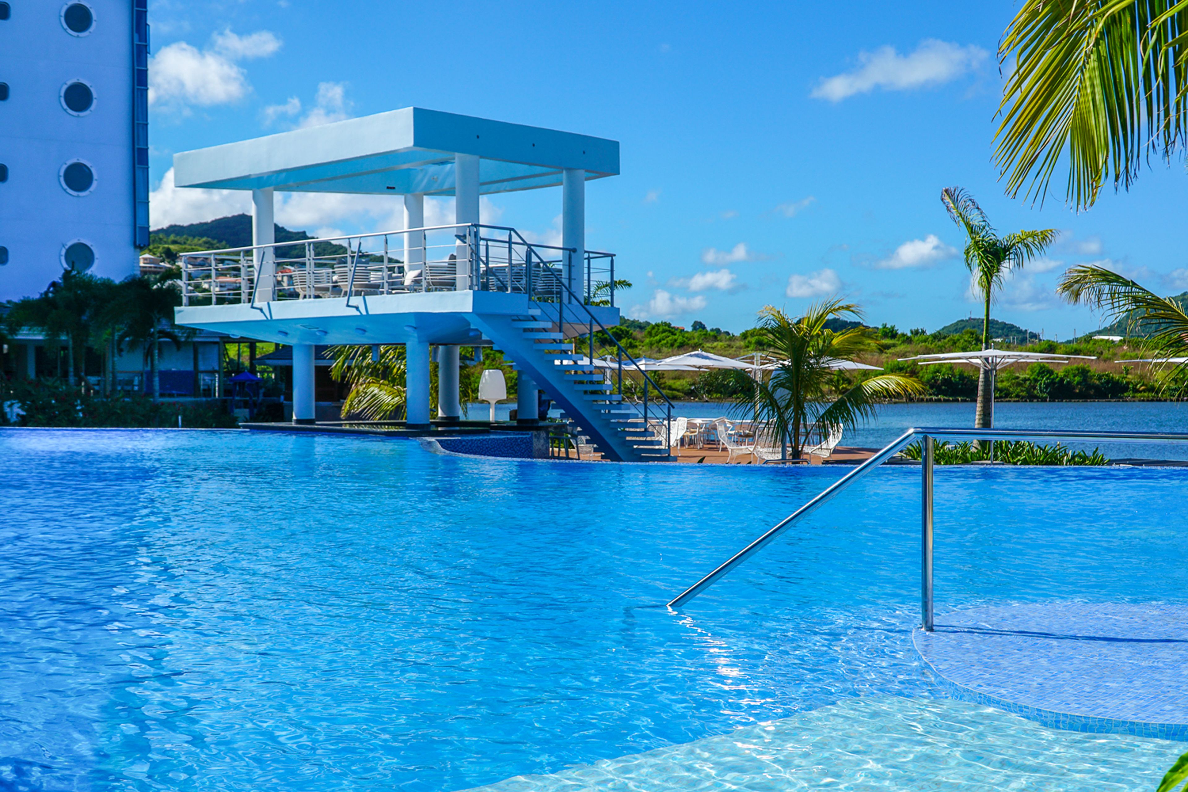 Una piscina azul cristalina con una plataforma de clavados y escaleras, rodeada de palmeras y un edificio con fachada blanca.