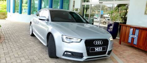 Un Audi plateado con la placa 1453 estacionado frente a un edificio con ventanas de marco azul y una entrada empedrada con ladrillos.
