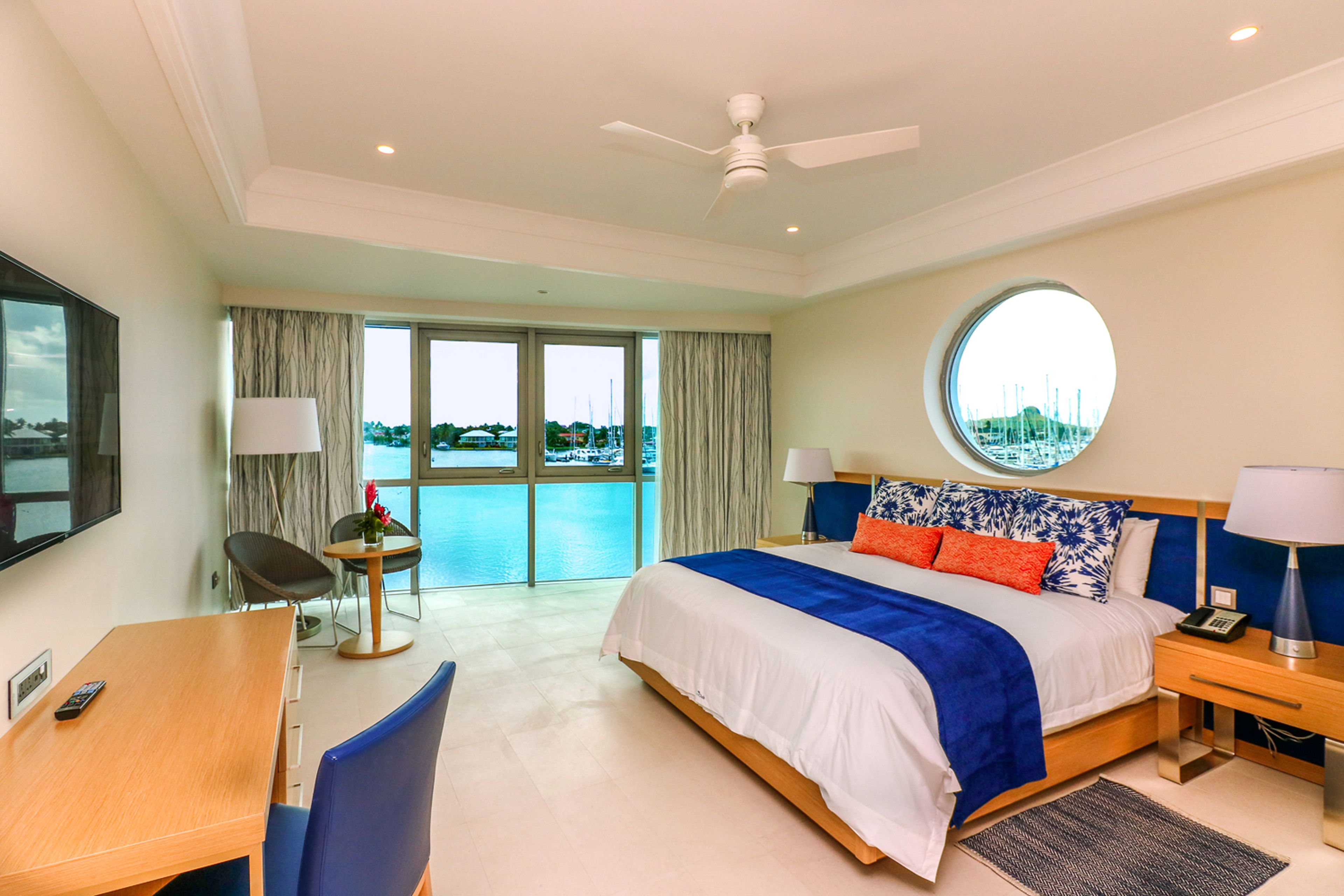 A hotel room with a bed, a desk, a chair, a round window, a ceiling fan, and a view of the sea.