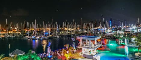 A marina at night with boats docked, a swimming pool area with lounge chairs, and a small building with a balcony.