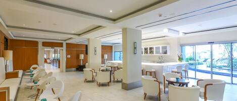A spacious hotel lobby with white chairs and tables, a reception desk, and large windows offering a view of the outdoors.