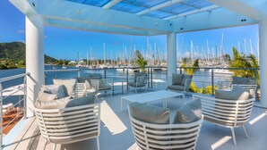 Un espace extérieur aménagé avec des meubles blancs, abrité et offrant une vue sur une marina où sont amarrés de nombreux bateaux.