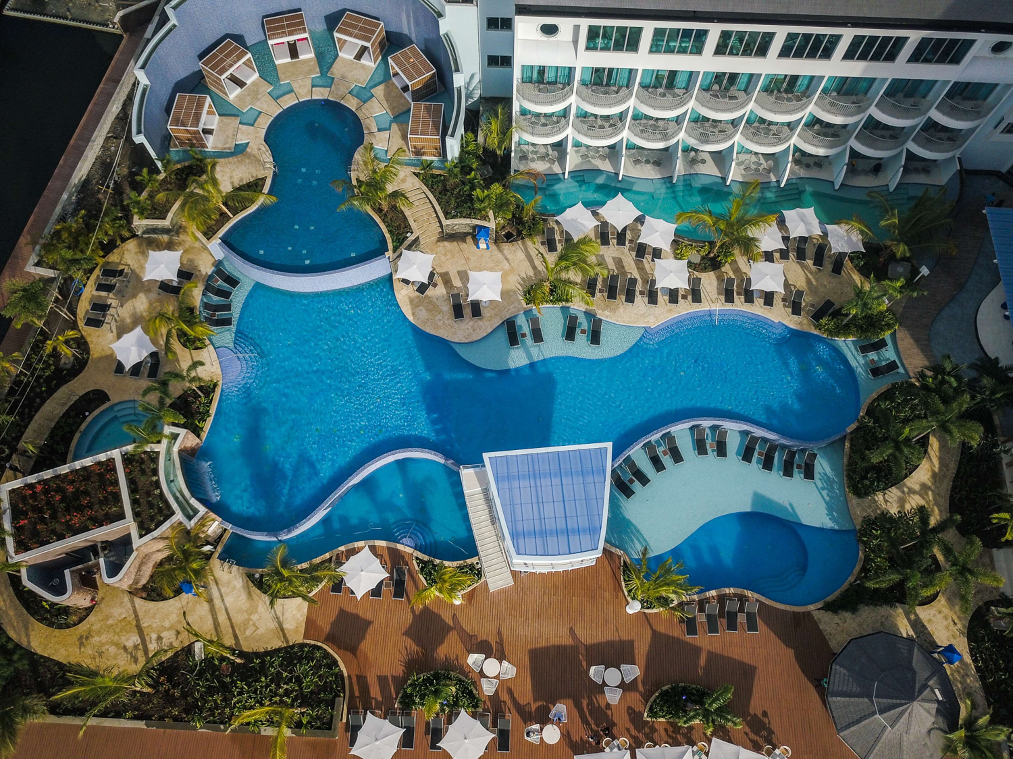 Eine Vogelperspektive zeigt einen luxuriösen Ferienort mit einem großen zentralen Pool, zahlreichen Liegestühlen und einem Gebäude mit Balkonen.